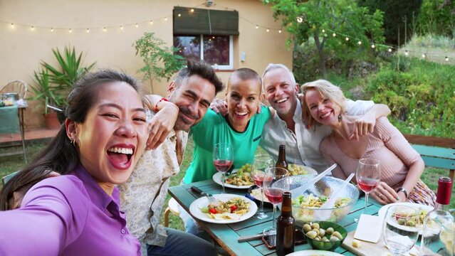 Happy asian woman taking a selfie photo using smartphone with diverse adult friends posing for the picture. Men and women celebrating a evening dinner party reunion together. High quality 4k
