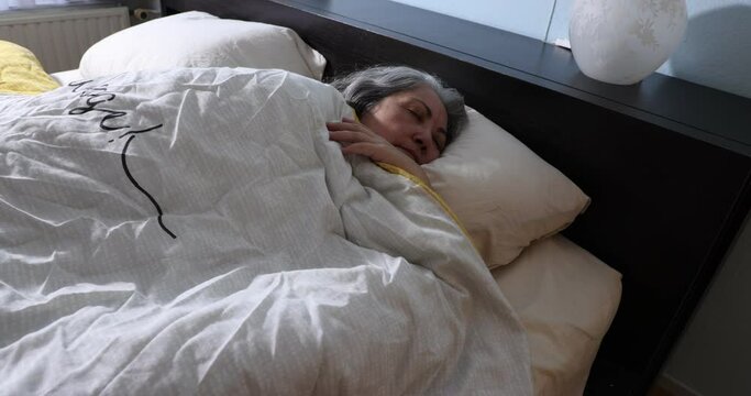 Mature woman sleeping peacefully without wanting to wake up to continue dreaming, sunlight coming through the window, moving slowly, long gray hair, white duvet, absolute tranquility