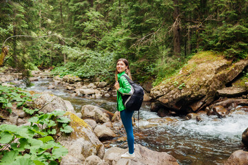 Naklejka na ściany i meble Traveler hiker with backpack enjoys landscape of mountain river in Carpathian forest. Young woman walks on rocks