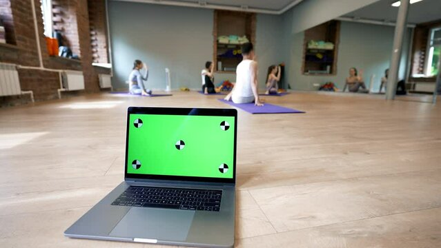 Open laptop with green chromakey screen against people doing yoga exercise on mats. Modern device stands on parquet in big hall closeup