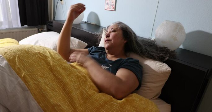 Mature woman just woke up and sits calmly in bed, sunlight coming through the window, moving slowly, long gray hair, yellow duvet, smiling to start a new day
