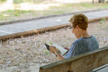 Rear view, portrait. Senior woman sitting and relaxing on the park bench, reading a book.