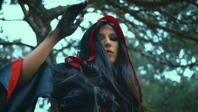 Fantasy portrait woman witch dancing a ritual dance. Sexy girl vampire magician shakes head hand, long hair hood on head slow motion. Black red dress, art costume. Tree green leaves forest, blue sky