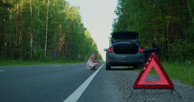 The car of a young girl broke down on the road, she put up an emergency sign, looked at the breakdown and took out the tools from the trunk.