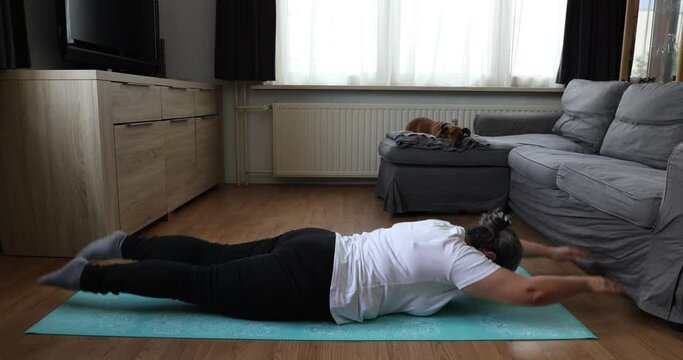 Senior Woman Doing Upside Down Exercises To Strengthen Waist On Yoga Mat, Raising Her Legs And Arms, Her Dog In Background. Concept Of Exercises For Mature People At Home And Healthy Lifestyle