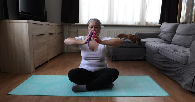 Senior woman sitting on yoga mat using chest strengthening device, her dachshund dog yawning in background. Concept of exercises for mature people at home and healthy lifestyle