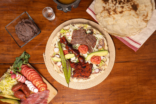Top view of Mexican food Tlayuda de cecina y chorizo and its preparation ingredients on a wooden table, frijoles, beans, cecina, sausage, lettuce, sauce
