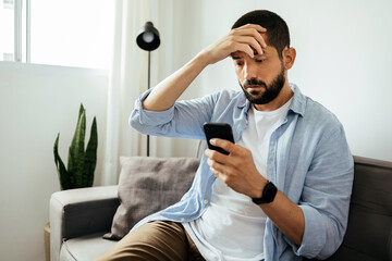 Sad man checking smartphone sitting on a sofa at home - Powered by Adobe