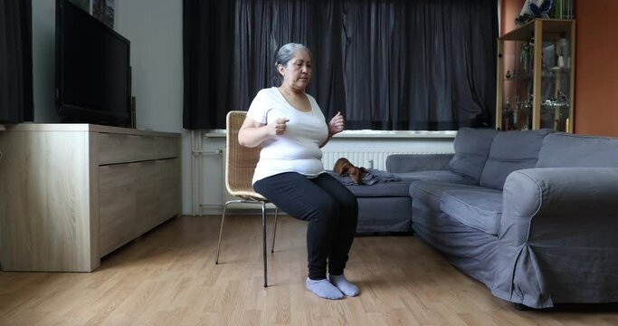 Senior woman stretching strengthening her legs and arms sitting on a chair, her dachshund dog in the background. Concept of exercises for mature people at home and healthy lifestyle