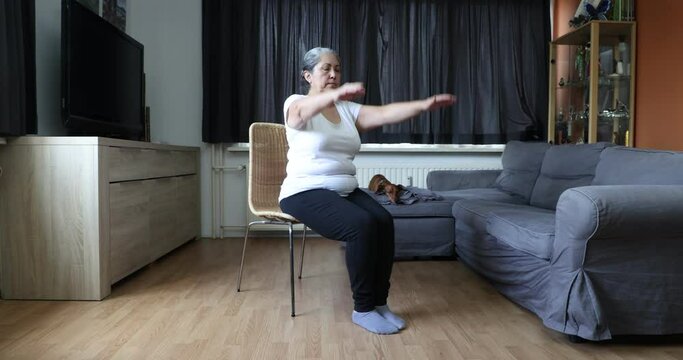 Senior woman doing arm stretch sitting on chair, her dachshund dog in background. Concept of exercises for the mature people at home and healthy lifestyle