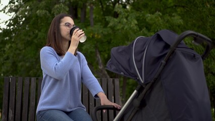 A young girl in glasses with a stroller in the park drinks coffee. Mom sits with a glass of coffee and shakes the stroller with a sleeping baby.