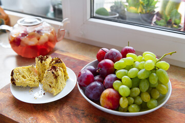 slices of homemade cake on a plate, green grape and plums, a glass teapot with stewed fruits on a...