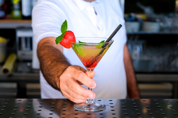 Hand of bartender serving fresh yellow, red summer fruit cocktail at bar counter on sunny day. Selected focus, shallow depth of field.