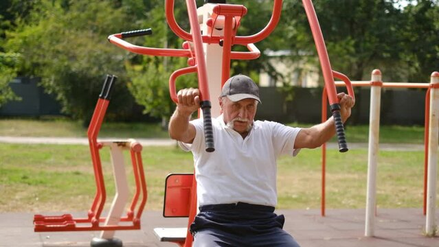 Senior Man In Sweatpants And A T-shirt Pumps Muscles, Does Fitness Exercises On An Iron Simulator Outdoors, Plays Sports On A Special Site, Leads A Healthy Lifestyle