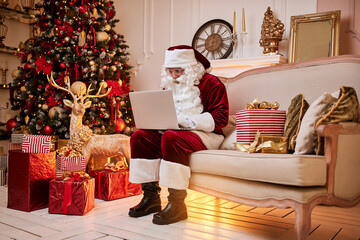 Santa Claus sitting at his home and reading email on laptop with сhristmas requesting or wish list near the fireplace and tree with gifts. New year and Merry Christmas , happy holidays concept