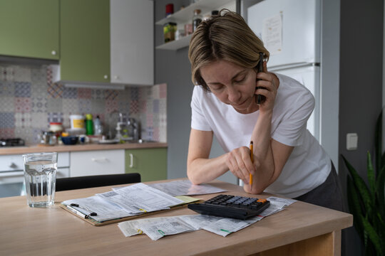 Mature woman talks on smartphone calling different hotlines. Middle aged female with upset expression tries to solve problems with unpaid bills checking papers on table. Utility debt and rates concept