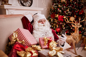 Santa Claus sitting on couch and talking on mobile phone near the fireplace and christmas tree with...