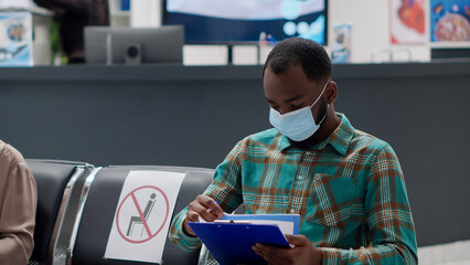 African american man filling in medical report to attend consultation appointment, waiting in facility reception lobby. Male patient with face mask sitting in waiting area during virus epidemic.