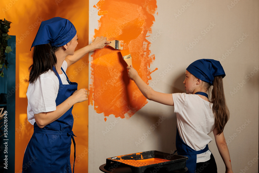 Wall mural Mom and kid using orange paint on walls and brush to renovate house interior, painting apartment room together. People having fun with housework redecoration, diy decorating job.