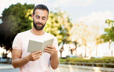Fototapeta premium Handsome smiling latin man reading book in park, copy space. Pensive student studying, education concept 