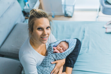 Pretty caucasian married woman holding a newborn baby boy in her arms, sitting on a bed in a bedroom. High quality photo