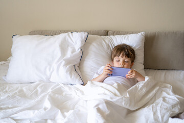 Happy little boy talking on smartphone, looking aside while sitting on bed