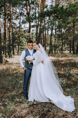 Beautiful newlyweds are hugging in the forest. Wedding portrait, photo of a stylish groom and a cute brunette bride in a white long dress with a bouquet of roses.