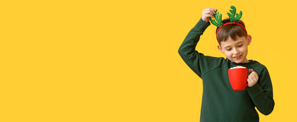 Funny little boy with reindeer horns and hot chocolate on yellow background with space for text