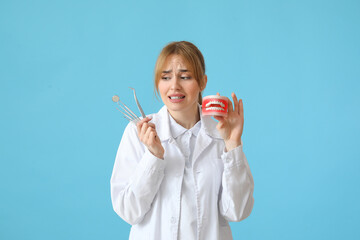 Scared female dentist with plastic jaw model and tools on blue background