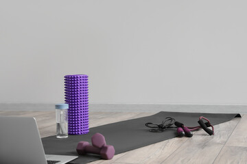 Foam roller, bottle of water, dumbbells and laptop on mat near grey wall