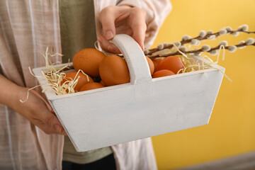 Woman holding basket with chicken eggs and pussy willow branches on color background, closeup