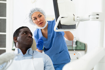 Qualified asian female dentist pointing at monitor explaining diagnosis to focused african american...
