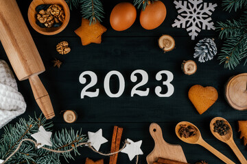 White wooden numbers 2023 lying on black wooden table with branches of Christmas tree, baking accessories and ingredients. Merry Christmas and Happy New Year 2023. Top view