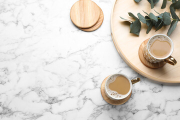 Mugs of coffee, stylish wooden cup coasters and eucalyptus branches on white marble table, flat...