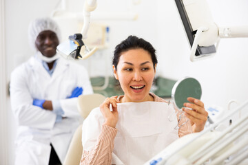 Satisfied asian woman looking in the mirror at the cured teeth at the dentist