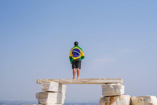 A faraway view of a man standing on a huge pile of flat rocks, looking to Horizon with brazilian Flag.