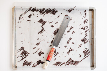 Spatula with scraps of melted, tempered semi-sweet cocolate drizzle decoration on sheet of white baking paper - 530702549