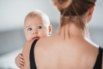 Portrait of sweet baby resting in mothers arms, looking at camera, touching mama shoulder. New mom holding little kid, embracing child with tenderness, love, care. Motherhood concept