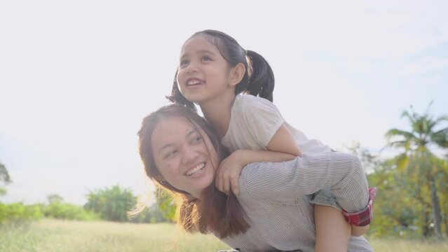 Adorable Asian kid ride on young mother shoulder or back spent time together in garden at sunset, happy family maternity playing, running outdoor, healthy mother piggyback daughter ride in park
