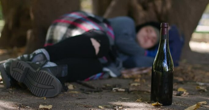 Close-up wine bottle with blurred homeless woman sleeping under tree at background. Caucasian alcoholic person napping outdoors on sunny summer day in slow motion. Cinema 4k ProRes HQ
