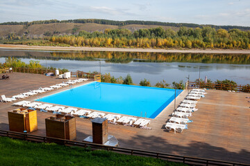 Obraz premium Blue pool without people on the riverbank in autumn. A great place to relax and unwind in nature. The pool and sun beds are waiting for visitors