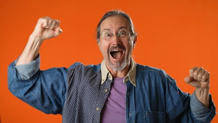 Portrait of excited jubilant overjoyed elderly bearded man 50s 60s doing winner gesture celebrate clenching fists say yes isolated on solid orange background studio