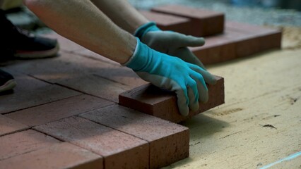 Low angle closeup of person installing brick pavers onto sand base wearing blue gloves.