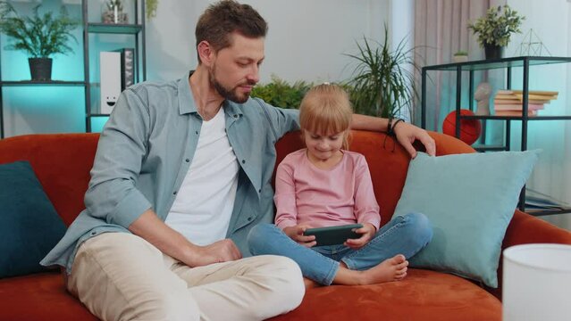 Happy family father or nanny and child kid daughter laugh use smartphone app look at screen play online game, daddy with child take selfie watch cartoons make video call on mobile sit on sofa at home