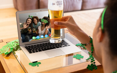 Naklejka na ściany i meble Mixed race woman holding beer having st patrick's day video call with friends on laptop at home