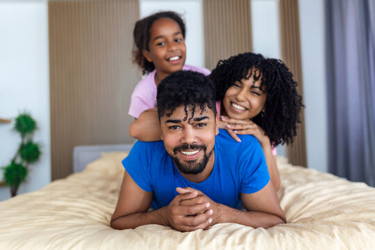 Portrait of beautiful young mother, father and their daughter looking at camera and smiling while lying on bed leaning on each other