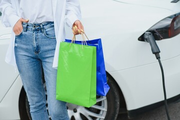 Happy young woman standing on city parking near electric car, charging automobile battery from small city station, holding shopping bags.