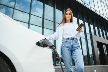 Phone in hands. Woman on the electric cars charge station at daytime. Brand new vehicle.