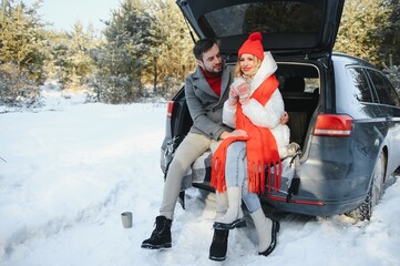 Couple in love sitting in car trunk drinking hot tea in snowy winter forest and chatting. People...