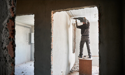 Professional strong destroyer standing on desk near gray brick wall covered with plaster and breaking it using jackhammer. Redevelopment of apartment in order to expand living space.